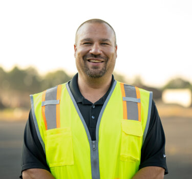 A man in a neon yellow safety vest smiling outdoors with a blurred background.