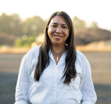 A woman in a white button-up shirt standing outdoors, smiling at the camera.