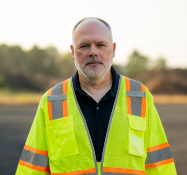 A man in a black polo shirt and high-visibility vest standing outdoors, looking at the camera.