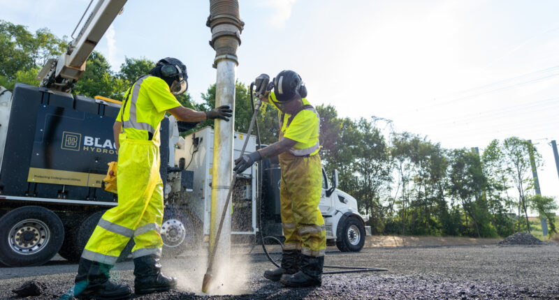 Two workers in yellow safety suits operating a vacuum excavation system, with dust and debris in the air.