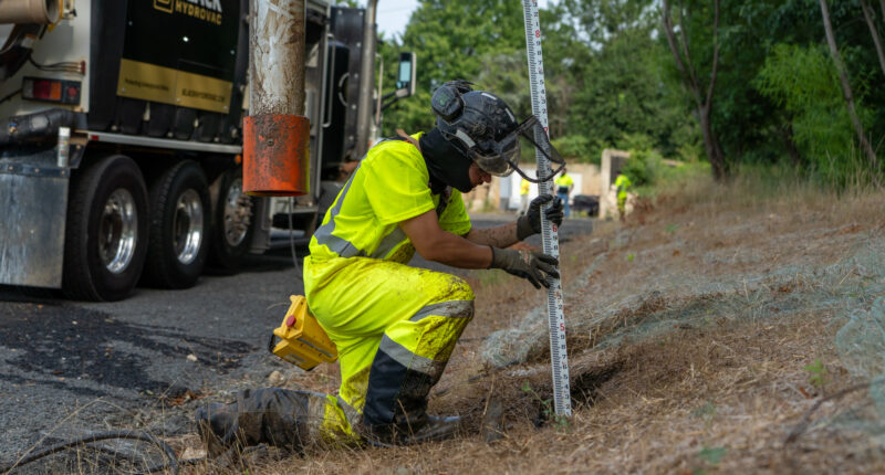 A construction worker in yellow safety gear using a measuring tool on a cut hole.