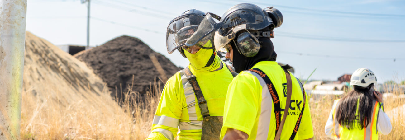 Two construction workers in safety gear having a discussion near tall grass.