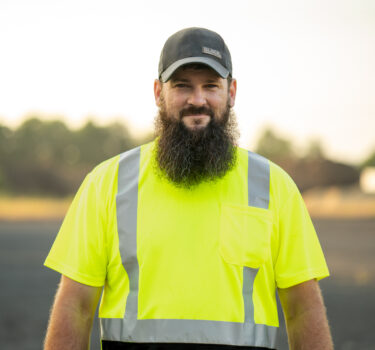A bearded construction worker wearing a neon yellow safety shirt and black cap, standing outdoors.