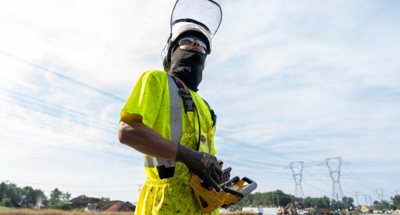 A construction worker in a bright yellow safety uniform and helmet, standing near power lines.