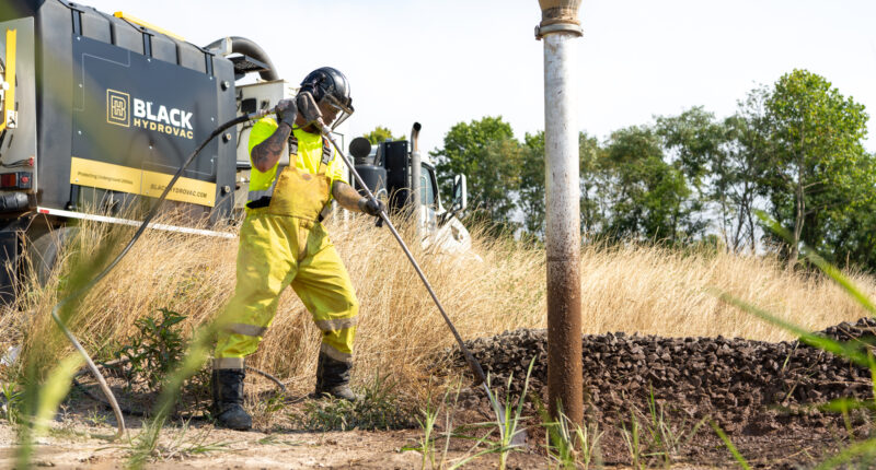 A worker in a yellow uniform using a tool near an overgrown roadside area.